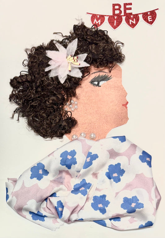 This is a handmade card of a woman named Lady Flora. Her curly hair is styled with a soft pink flower clip, and she wears pearl-like earrings and a matching necklace. Her outfit is made from silky fabric with a pattern of blue and white flowers on a pale pink background. Her face is drawn in profile with gentle features and red lips. At the top corner, red glittery letters spell out “Be Mine.”