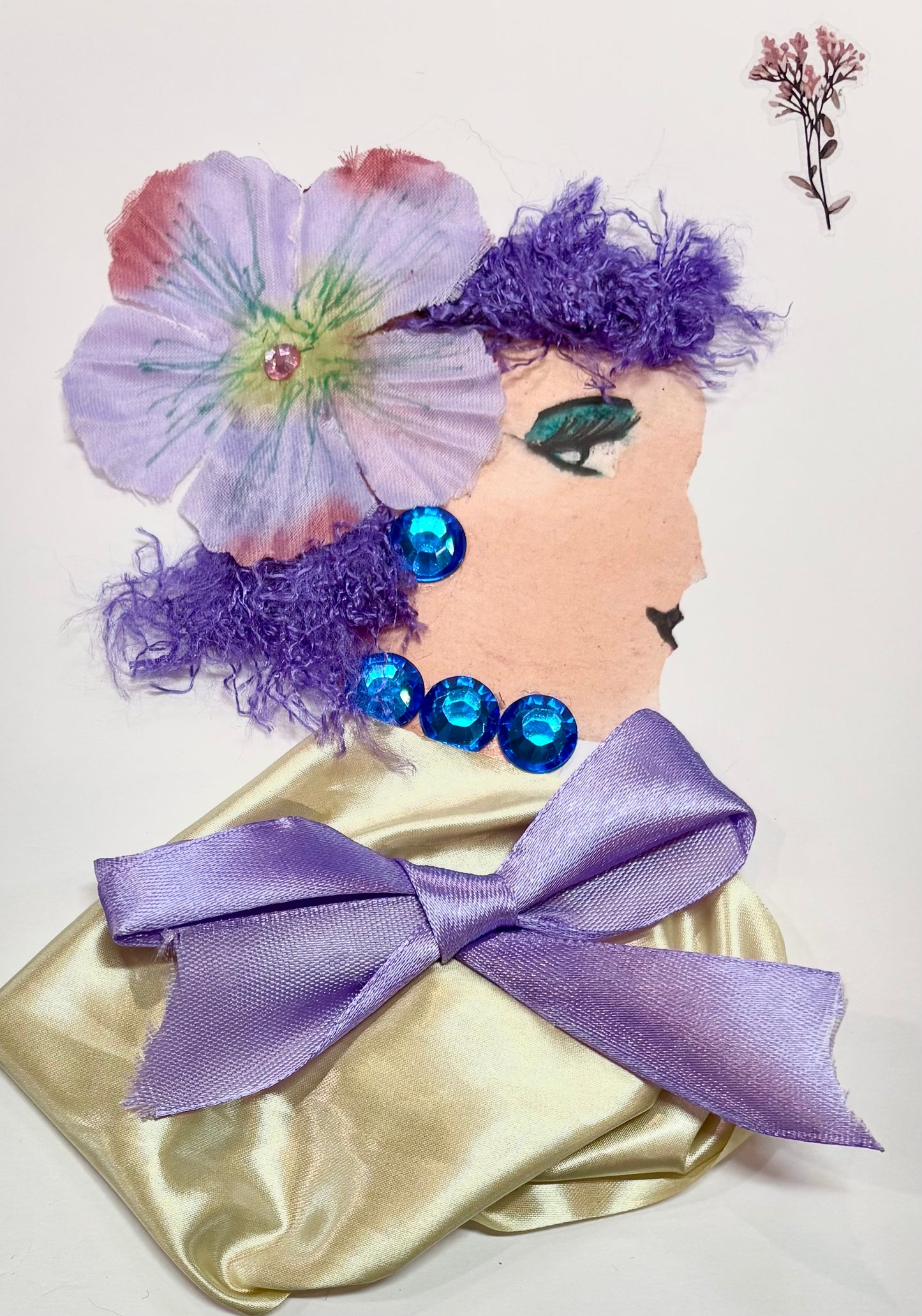 A woman has dark purple hair with a light purple flower as an accessory. She wears a gold blouse with a light purple bow placed in the center. She has sapphire blue gem earrings and a necklace. A dainty flower sits in the top right corner of the card. 