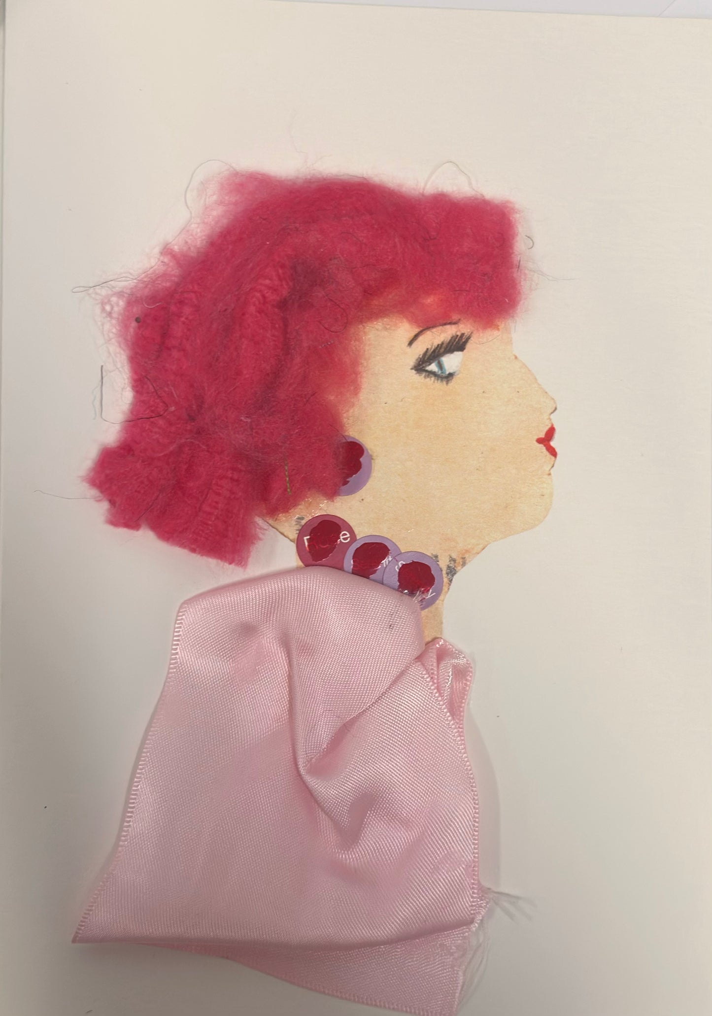 A handmade card shows a side-profile portrait of a woman with soft peach skin and bright, textured pink hair. She wears round red and purple jewel-like earrings and a matching choker, with a pale pink satin fabric draped around her shoulders. Her makeup includes bold eyelashes and red lips, giving her a stylish and expressive appearance.