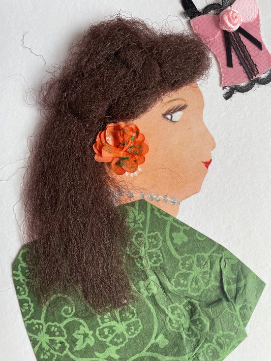 This is a handmade card of a woman named Luton Lily. She has thick, textured hair and wears a sparkly orange flower by her ear. Her necklace is a silver-toned embellishment, and she is dressed in green fabric with a floral pattern. Her face is drawn in profile with calm eyes and red lips. In the corner of the card, there's a small pink and black corset-shaped decoration with a tiny rose.