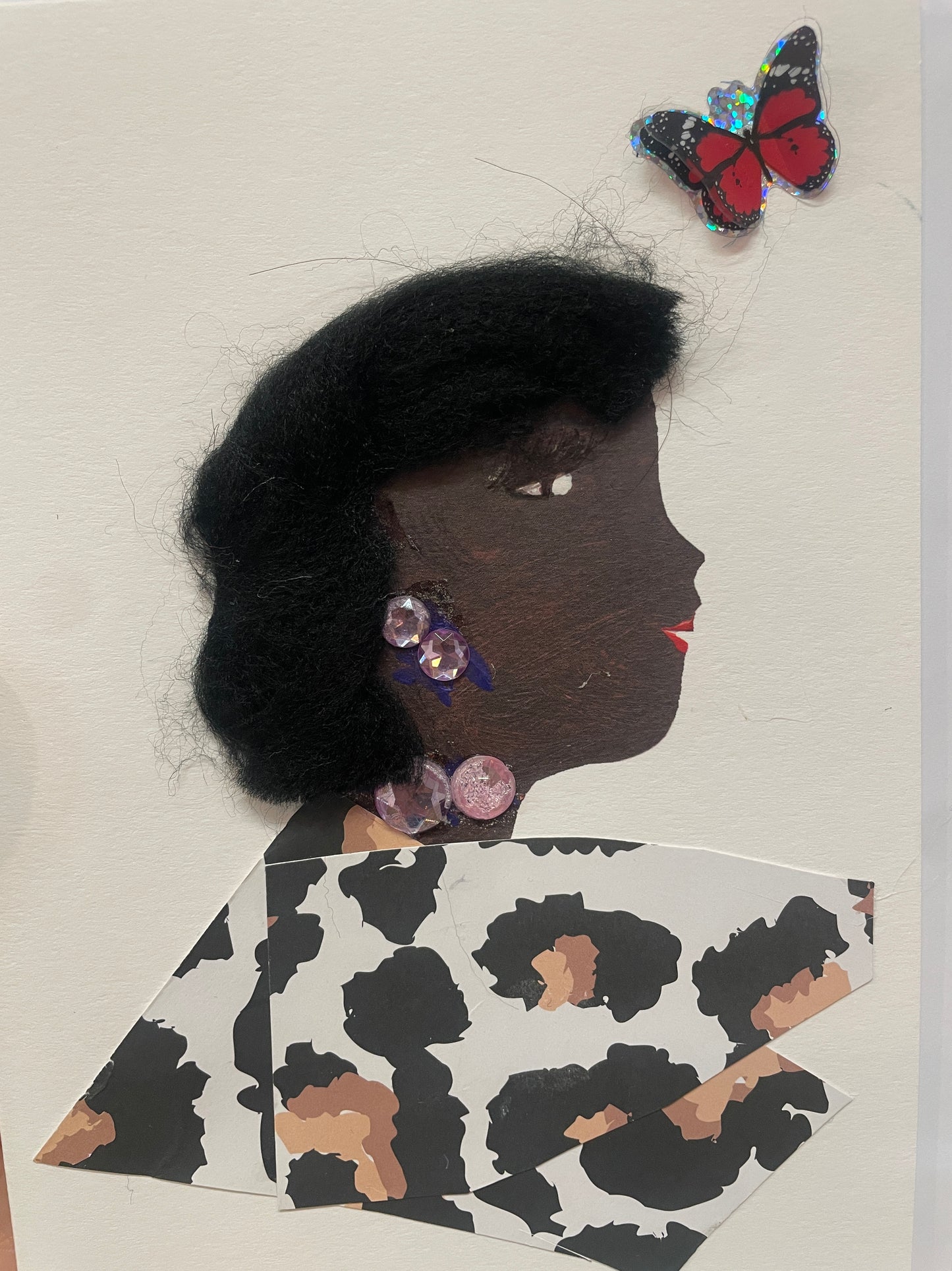  This is a handmade card of a woman named Mohak with black wool hair, purple earrings and necklace, and a cow-print outfit. A shiny butterfly sticker is nearby.