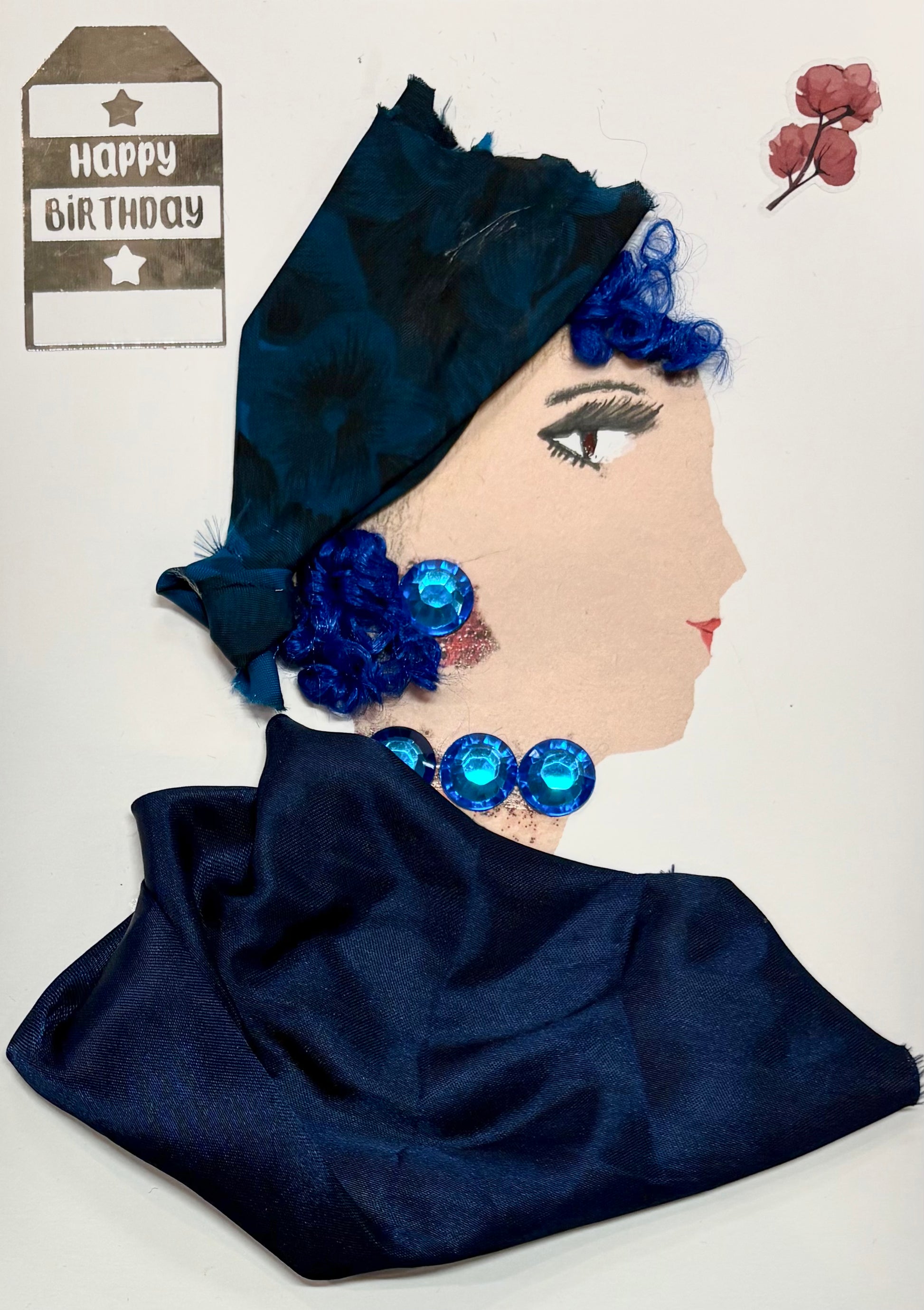 A woman has a deep blue headwrap with her curly blue hair peaking out. She has a dark blue blouse and wears blue gem earrings and a necklace. There is a "Happy Birthday" sticker in the top left and a dainty flower in the top right. 