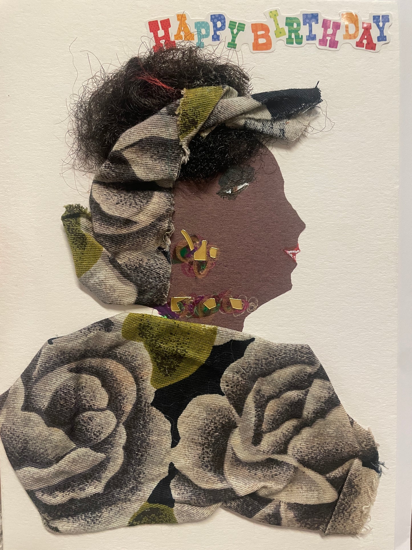 This is a handmade card of a woman named Temi Tope. Her hair is styled with black textured fibres and wrapped in floral fabric that matches her outfit. She wears a necklace and earring made of multicoloured gems. Her outfit is crafted from fabric featuring large grey roses with touches of green. At the top, colourful letters spell out “Happy Birthday.”
