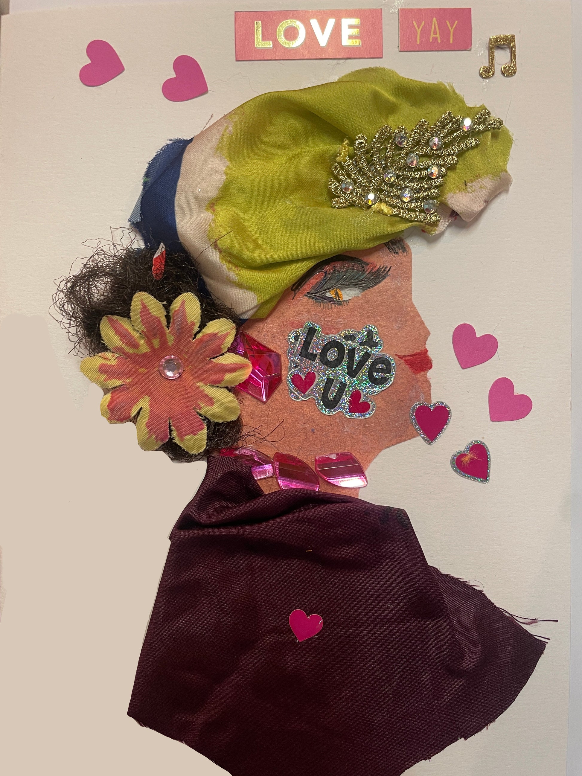 A handmade card of a woman in profile wearing a maroon dress, a yellow-green and navy head wrap with gold decoration, a pink flower on her shoulder, and surrounded by pink heart stickers and the word “LOVE”.