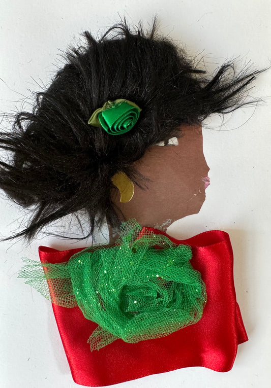 Handmade card showing the side profile of a woman with voluminous black textured hair, adorned with a small green ribbon rose. She wears a gold circular earring and pink lipstick. Her outfit features a red satin fabric with a green glittery netting rosette at the neckline. The background is plain white.