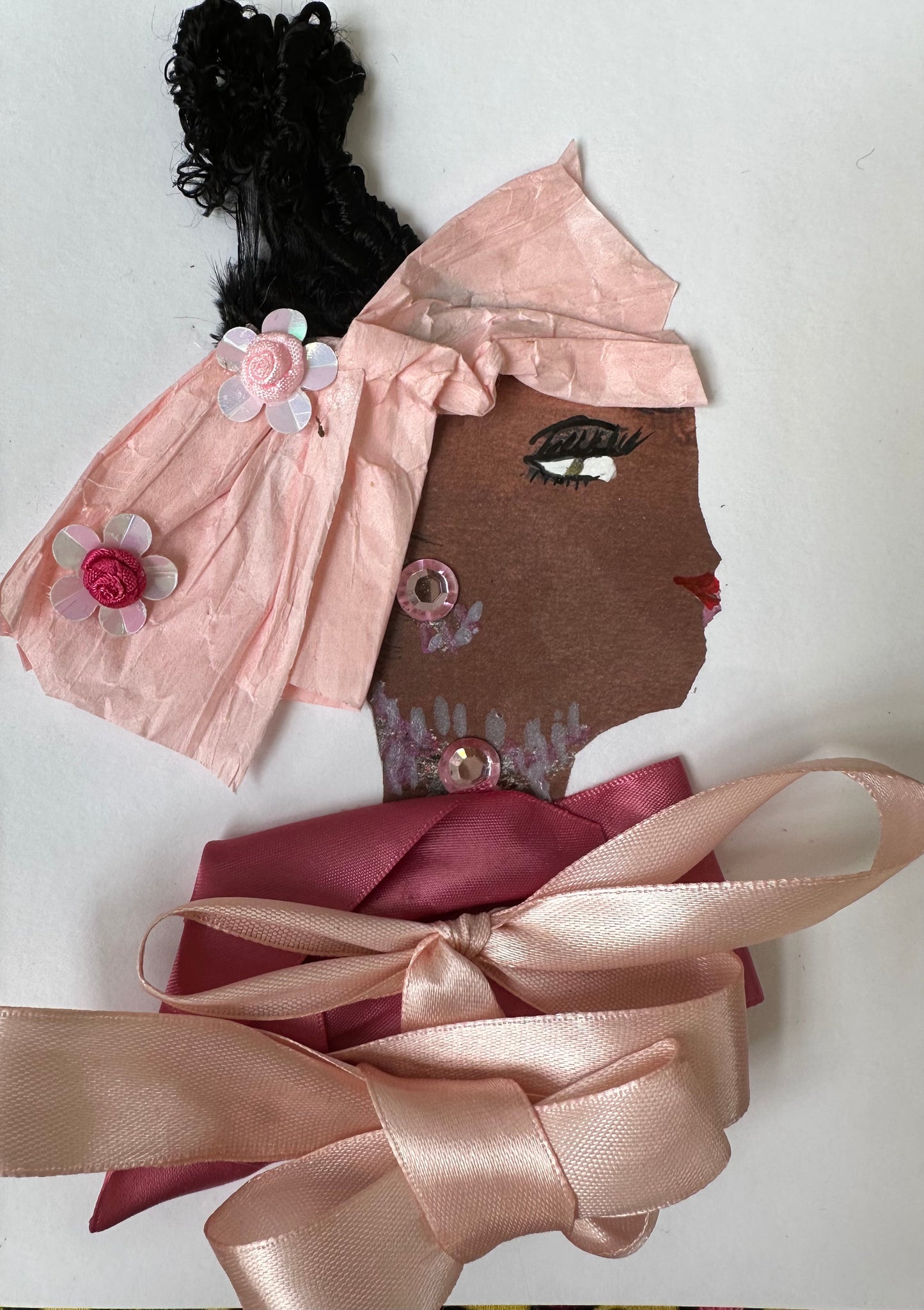 I designed this handmade card of a black woman dressed in a blouse that is composed of two light pink bows. Her hatinator is a light pink, bow-like shape with two flowers, and she has gemstones has jewellery. 