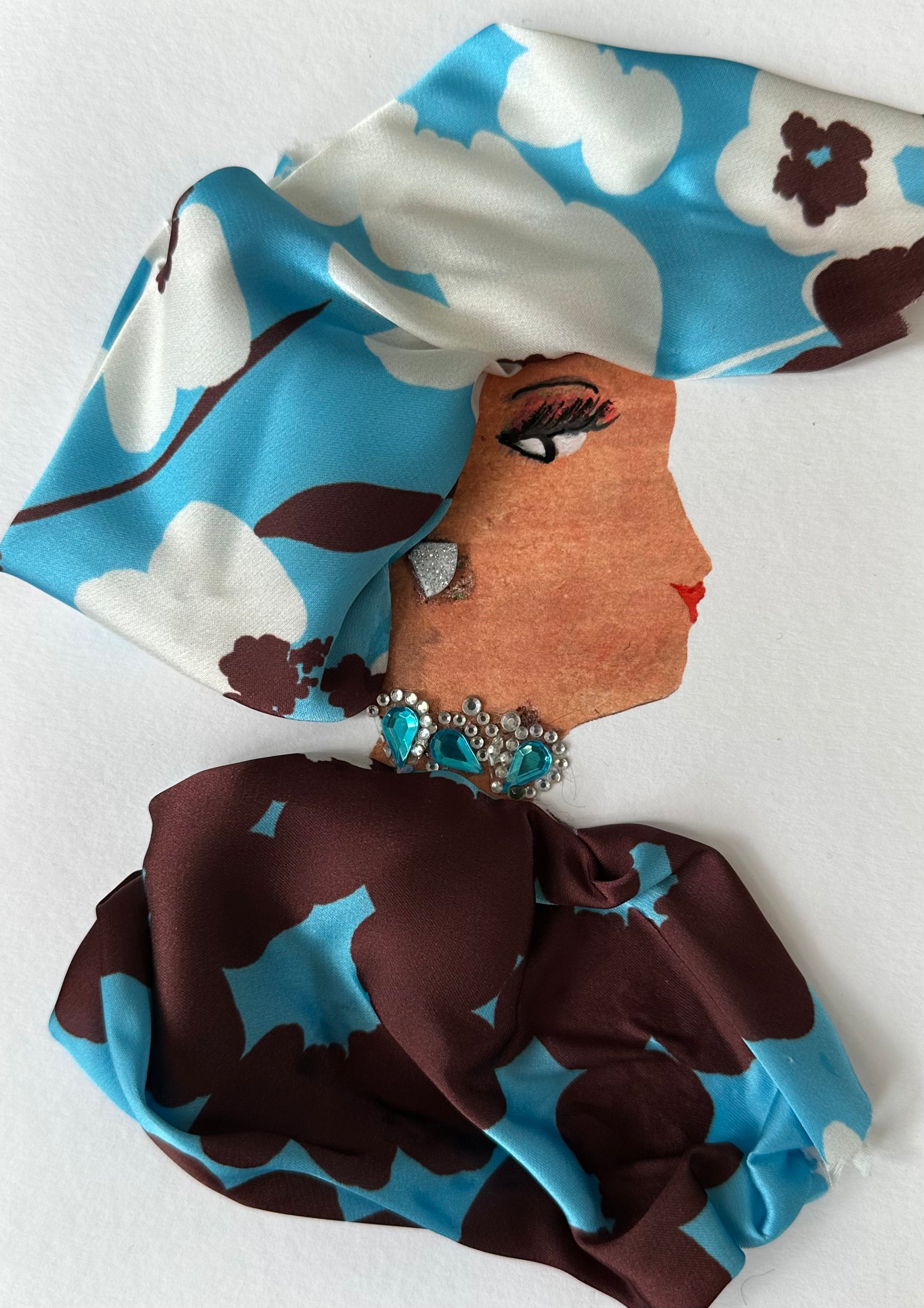 I designed this handmade card of a woman, Newbury Nadia, who is dressed in a powder blue, chocolate brown, and white floral material. This material makes up both her blouse and hatinator. Her outfit is completed with teal gemstone jewellery.