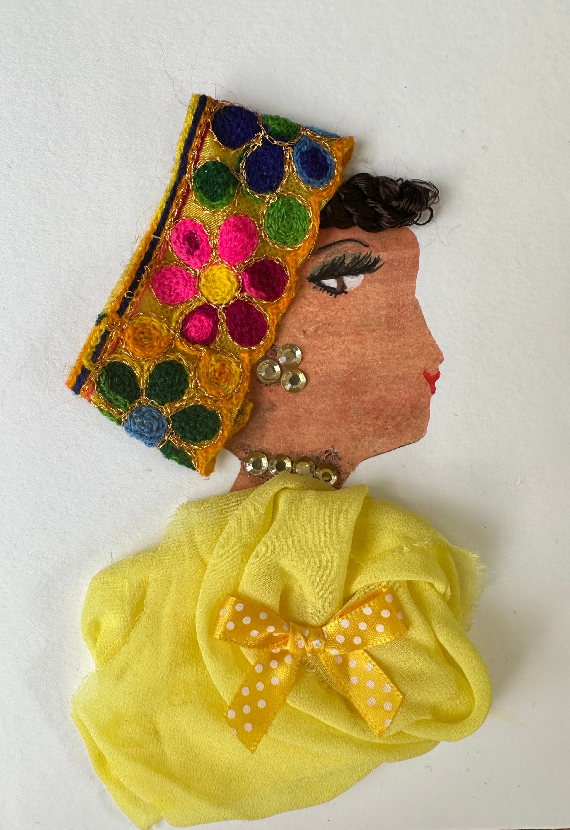 I designed this handmade card of a woman dressed in a yellow blouse that is pinned together with a yellow bow. She is wearing a headband with three flowers; one is green, one is pink, one is blue. The headband has a yellow background to match the blouse and touches her gemstone jewellery.