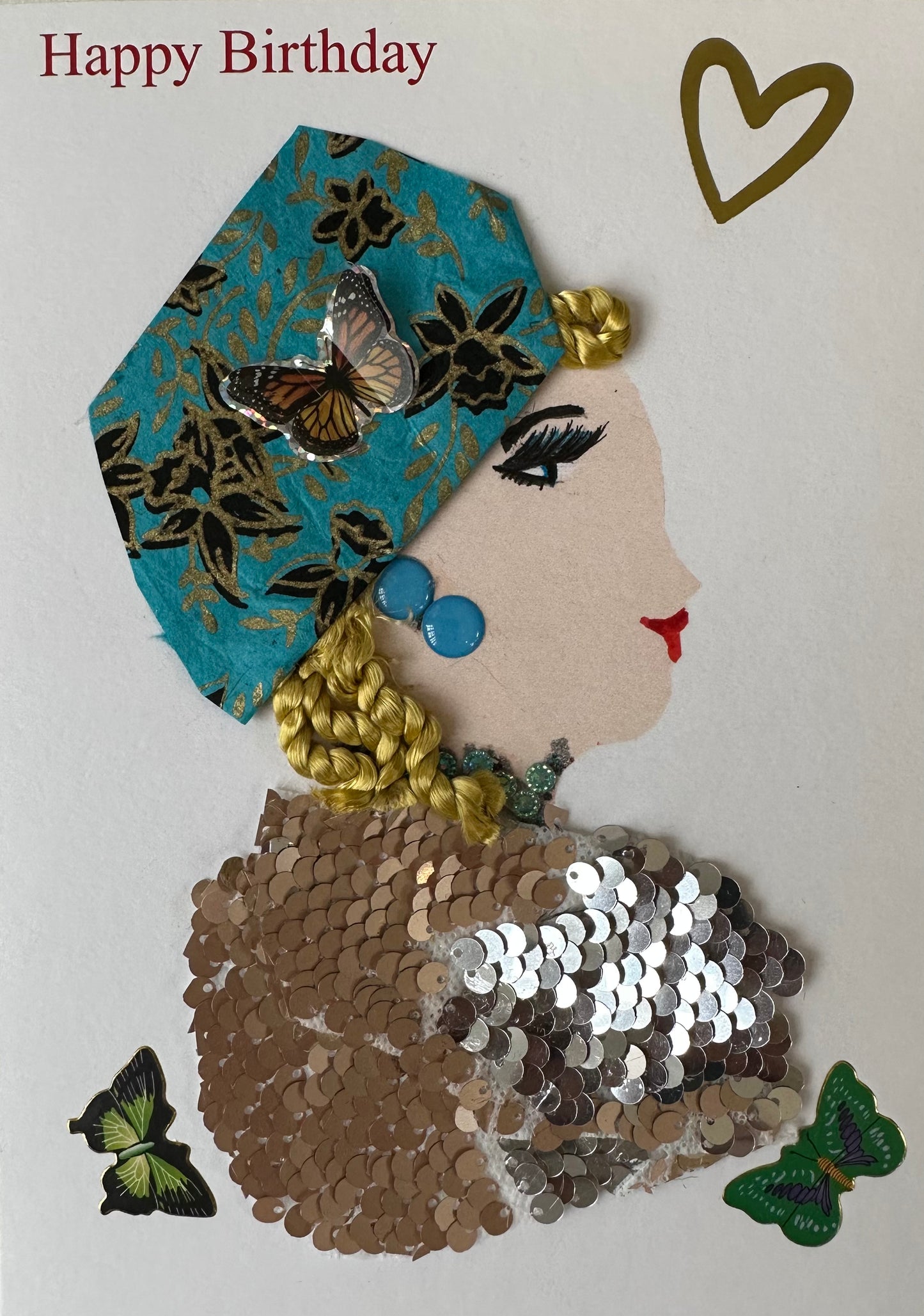 This handmade card is of a woman in a bronze coloured sequin top. Her blonde hair is covered by a teal coloured cloth with black and gold flowers. There is a heart in the upper right corner as well as "Happy Birthday" and butterflies.