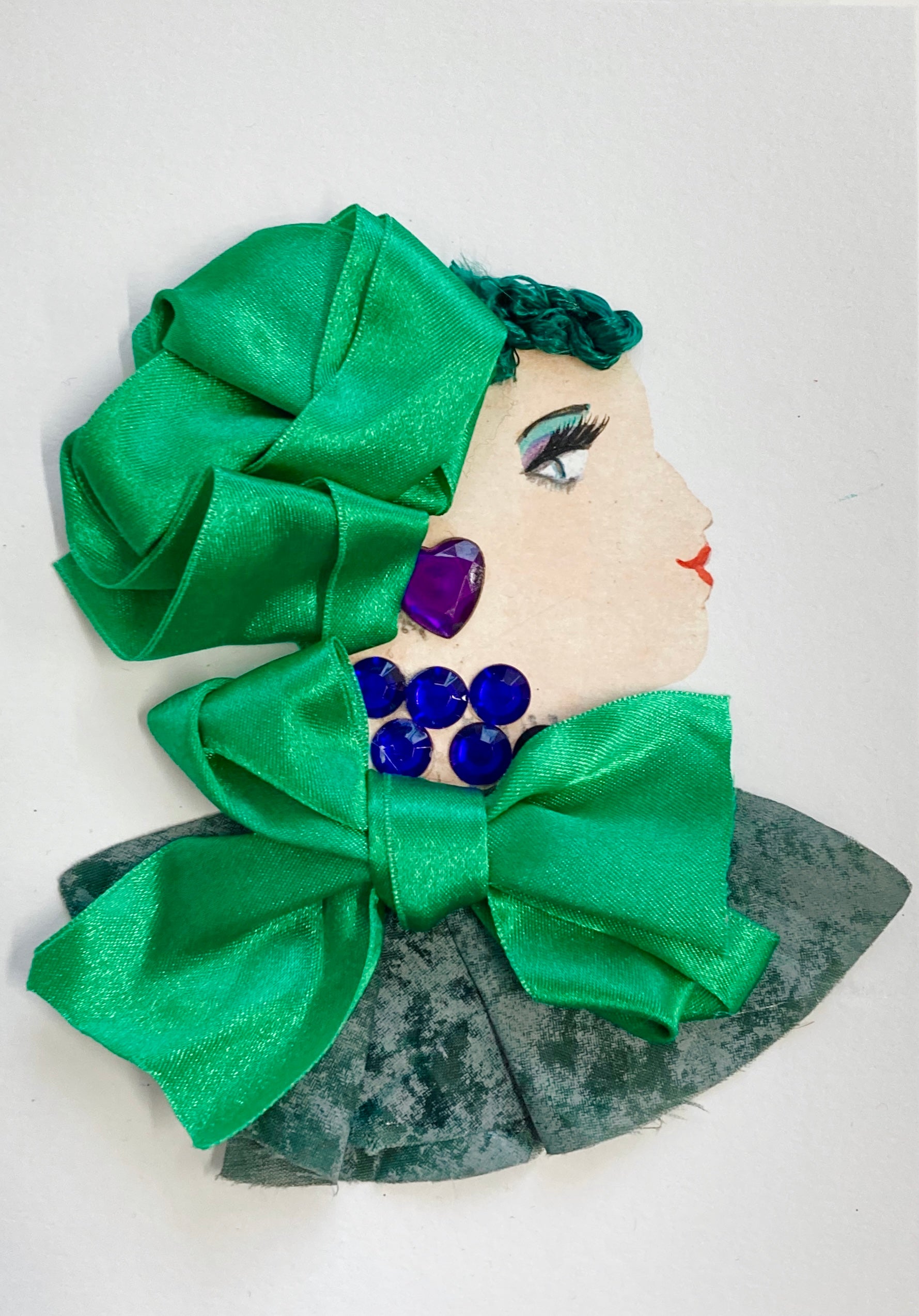 This card depicts a woman given the name Esmarelda. She wears a dark green textured blouse with a massive green bow at the neck. Her necklace is made of dark blue gems, and her earring is a purple heart gem. Her hair scarf is the same fabric as the bow on her dress. 
