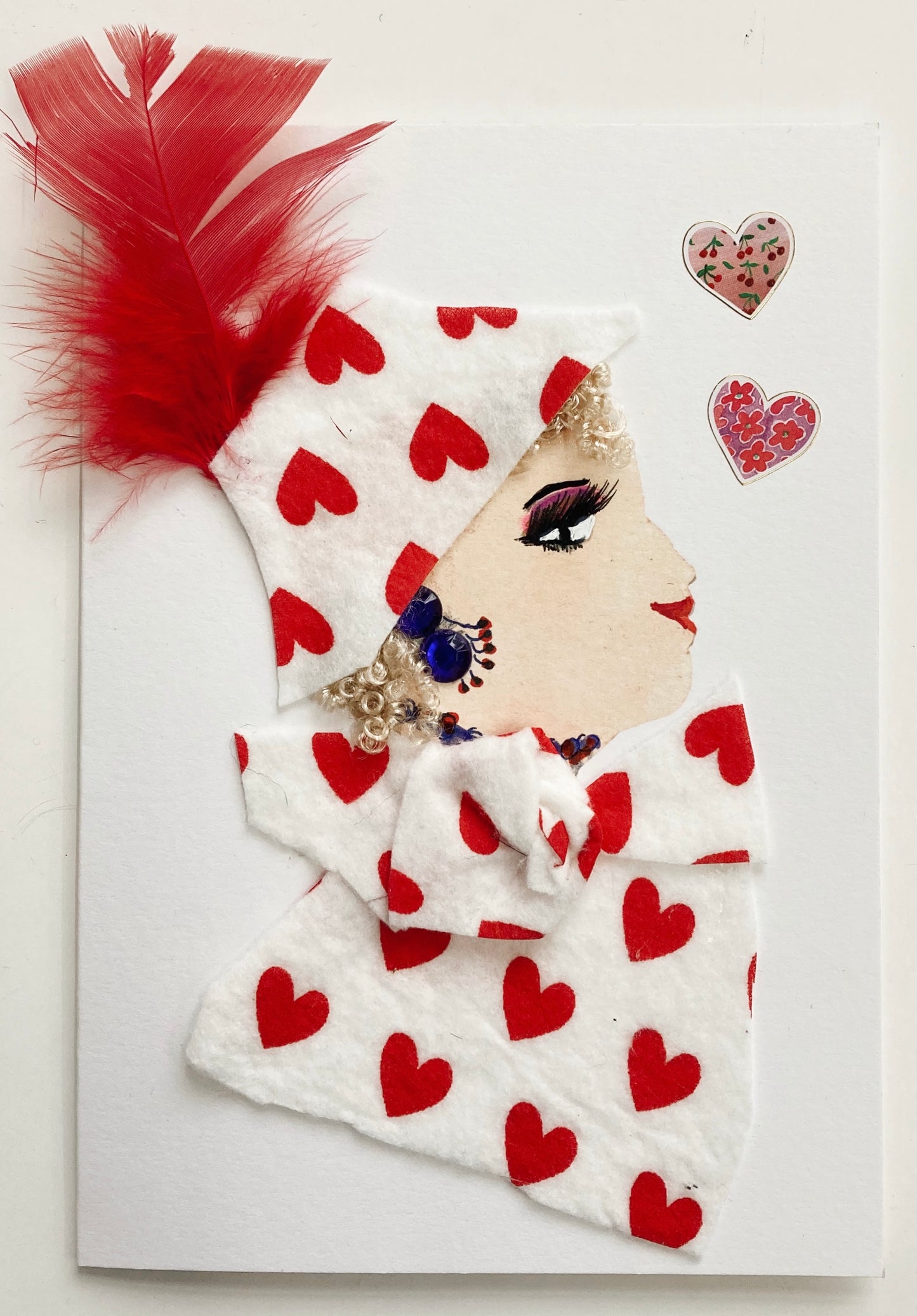 I designed this card of a woman who is named Liliana Liverpool. She has a white skin tone and wears a white hat with red hearts and a red feather coming out the top. She has blonde curls popping out the bottom of the hat. She wears a matching white blouse with read hearts all over. She wears navy blue gem jewellery. There are two hearts above her head that are both pink and red. 
