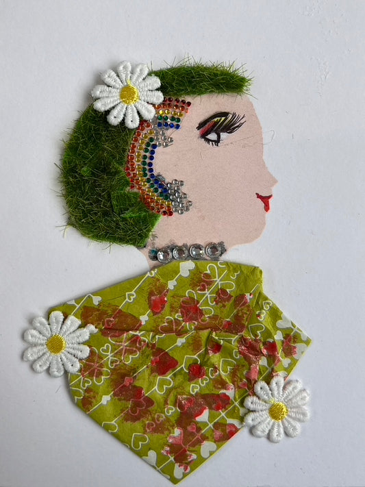 This card has been given the name Kaleidoscope Garden, and depicts a beautiful woman. Her hair is made of a grass material, has a small white daisy in it, and a rhinestone rainbow. Her blouse is green and has a red pattern on it. There are two more daisies at the bottom of the blouse. 