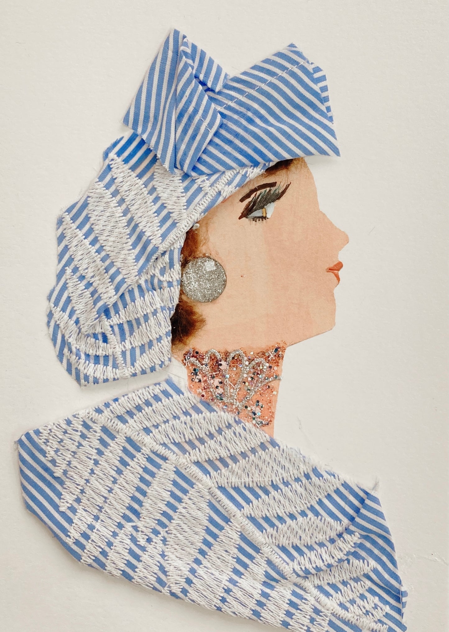 This card has been given the name Robin Redbridge. Robin wears a matching blouse and headscarf, both made of a blue and white pinstripe fabric with a leafy white embroidery overlaying. She wears a large, glittery gem for her earring and a glittery necklace. Visit my website for more 
