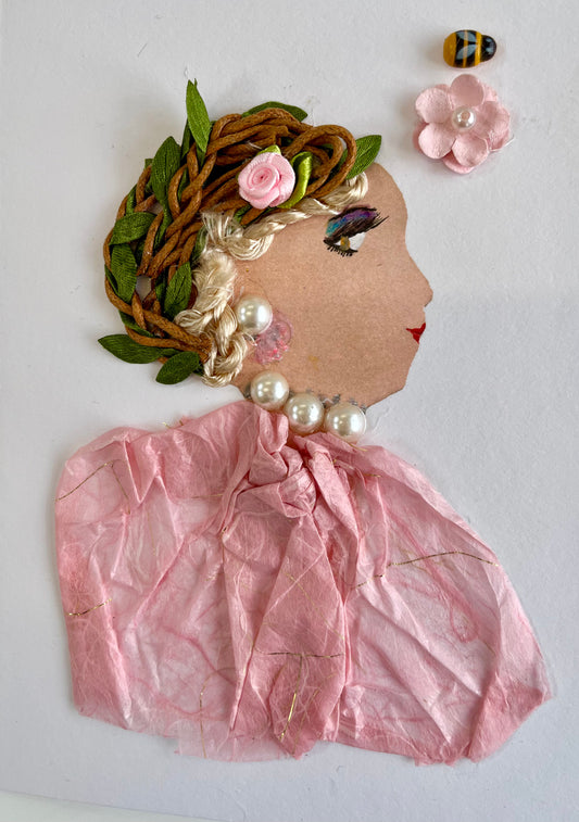 This card has been given the name Kendall. Kendall wears a baby pink paper material blouse. Her jewellery is white pearls. In her blonde hair is a twine and leaf headpiece that has a small baby pink rose in it. On the top right corner, there is a bumble bee and a baby pink daisy with a pearl at the center.