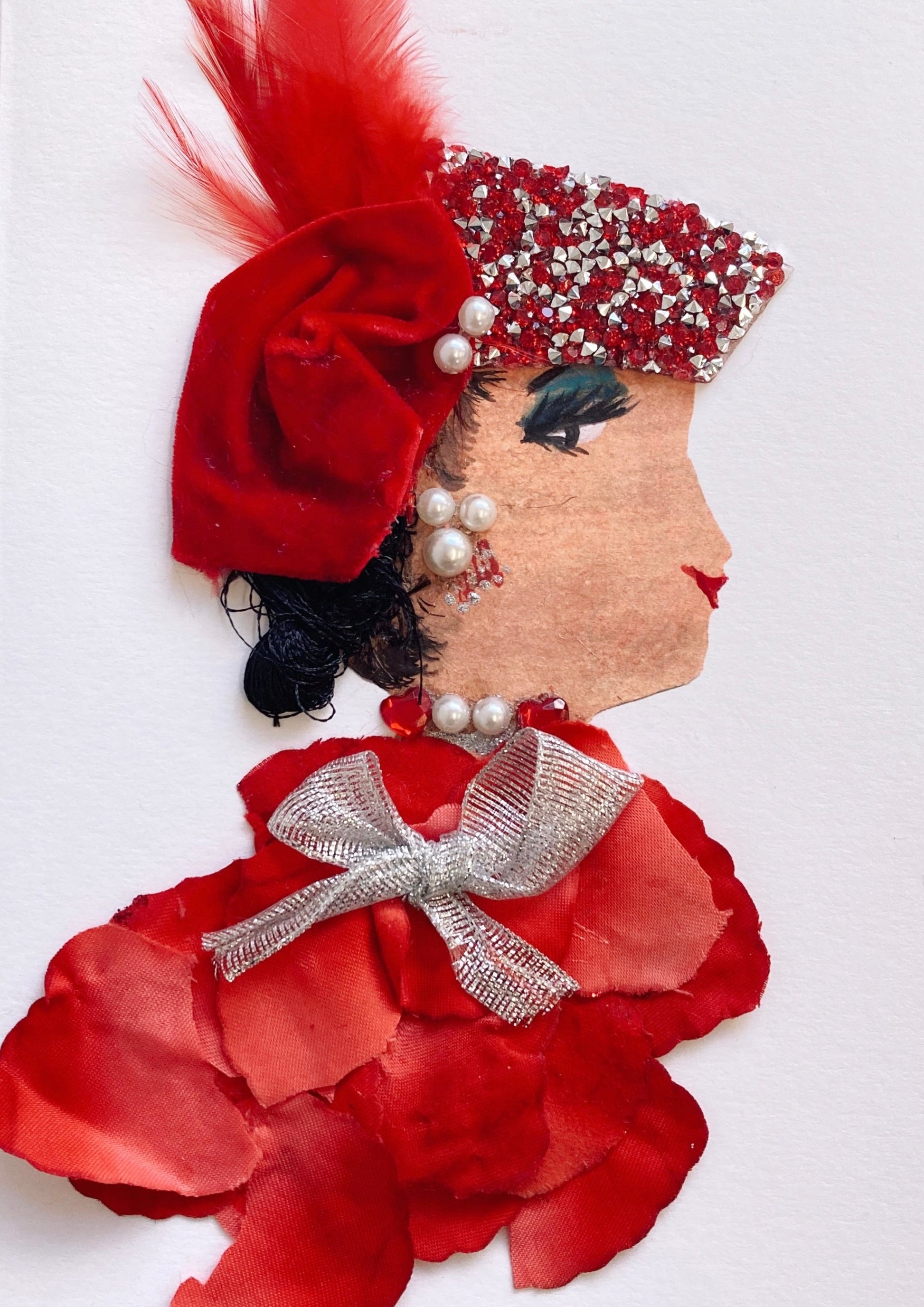 This card has been given the name Suzie. Suzie wears a red blouse made of fake flower petals, a silver bow around the neck, and a pearl necklace. She wears a headdress which has red and silver diamantés, a red feather, and some red velvet fabric. Her black curly hair peeks out of the bottom of the headdress. 