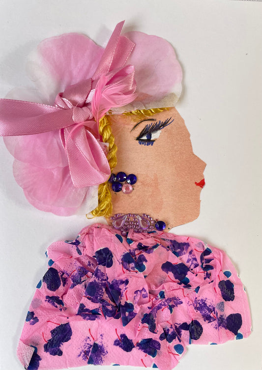 This handmade card called Caroline Colindale is adorned in a pale pink blouse, decorated by dark blue/purple brush strokes, partnered with a petal-adorned headband with a delicate pink ribbon atop her blonde tresses. Completing her ensemble are dusky purplish earrings.