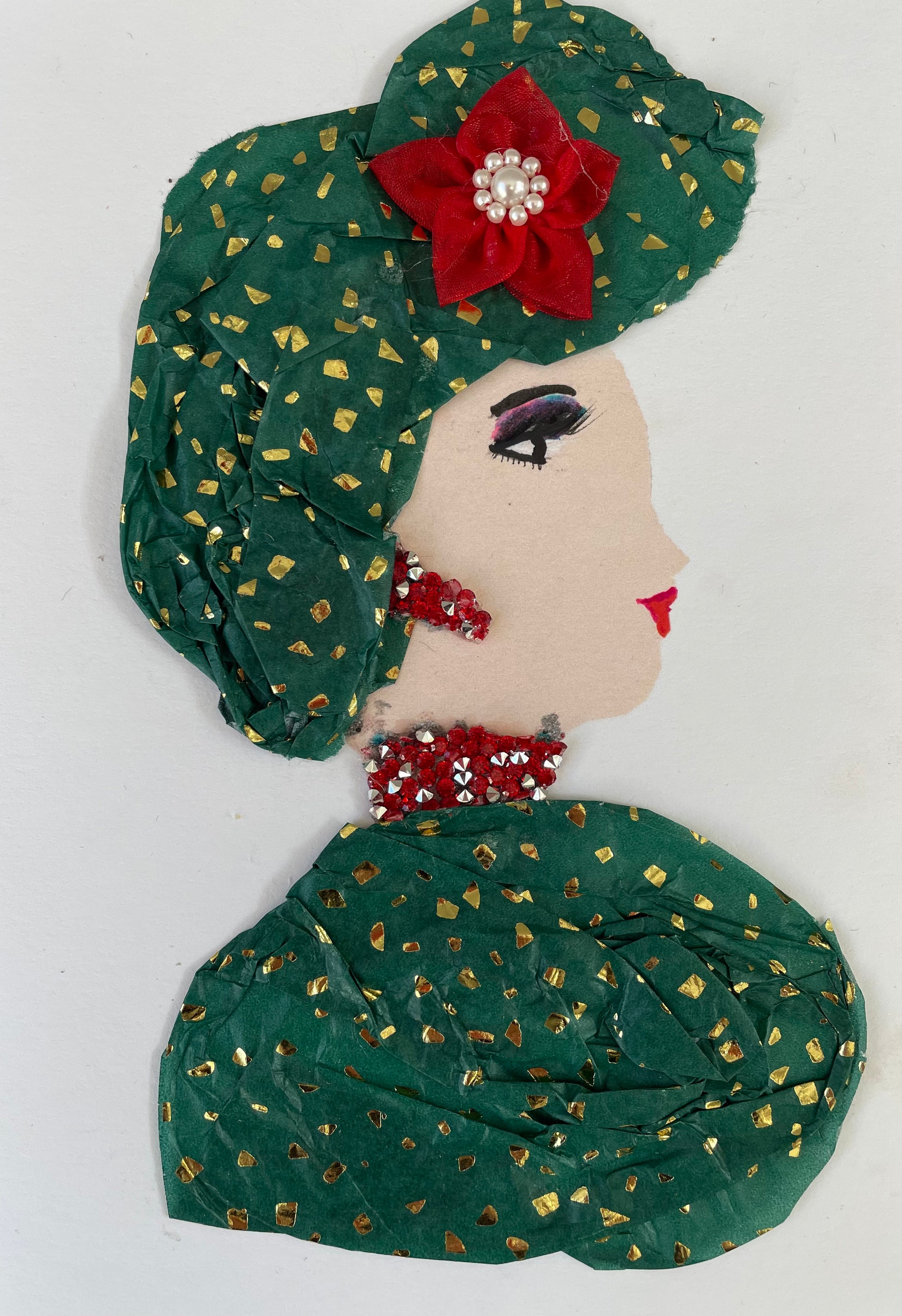I have called this card Norwood Nina. She is wearing a green blouse with gold patterned confetti on it. Her headpiece is the same print. She has matching earrings and a necklace that are red and silver bead-like pieces. She has a red flower in her headpiece