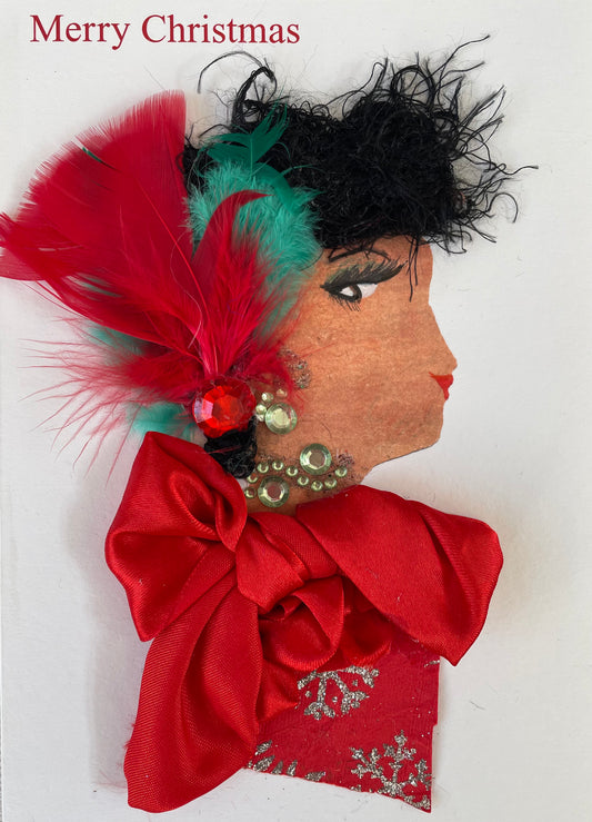 Handmade Christmas card featuring the side profile of a woman with curly black textured hair, decorated with red and green feathers and a red gem. She has red lips and green eye makeup. Her earring and neck accessory are made from green rhinestones. Her outfit is made of red fabric with silver snowflake patterns and is adorned with a large red satin bow. The background is plain white with “Merry Christmas” printed in red at the top left.
