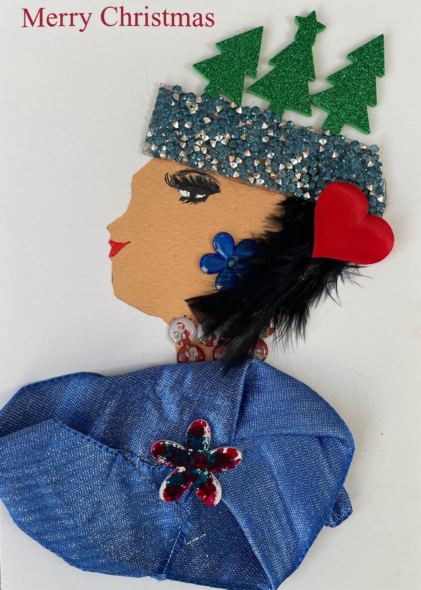This Christmas card is named Paulo Park. She  is wearing a soft coloured royal blue blouse with a red flower in the middle. She is wearing a silver and red necklace with royal blue earrings. She has a blue and silver diamanté headpiece on with 3 green trees on top. There is also a red heart in her hair. There is Merry Christmas written on top left in red. 
