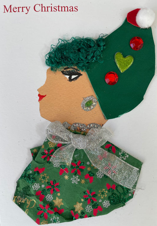 Handmade Christmas card featuring the side profile of a woman with green curly hair, a festive green hat decorated with red gems, a glittery green heart, and a white pom-pom at the tip. She has red lips and bold eye makeup. Her earring and neck accessory are silver and sparkly with green accents. She wears a green outfit with red and gold poinsettia patterns, topped with a large silver bow. The background is plain white with "Merry Christmas" printed in red at the top left.