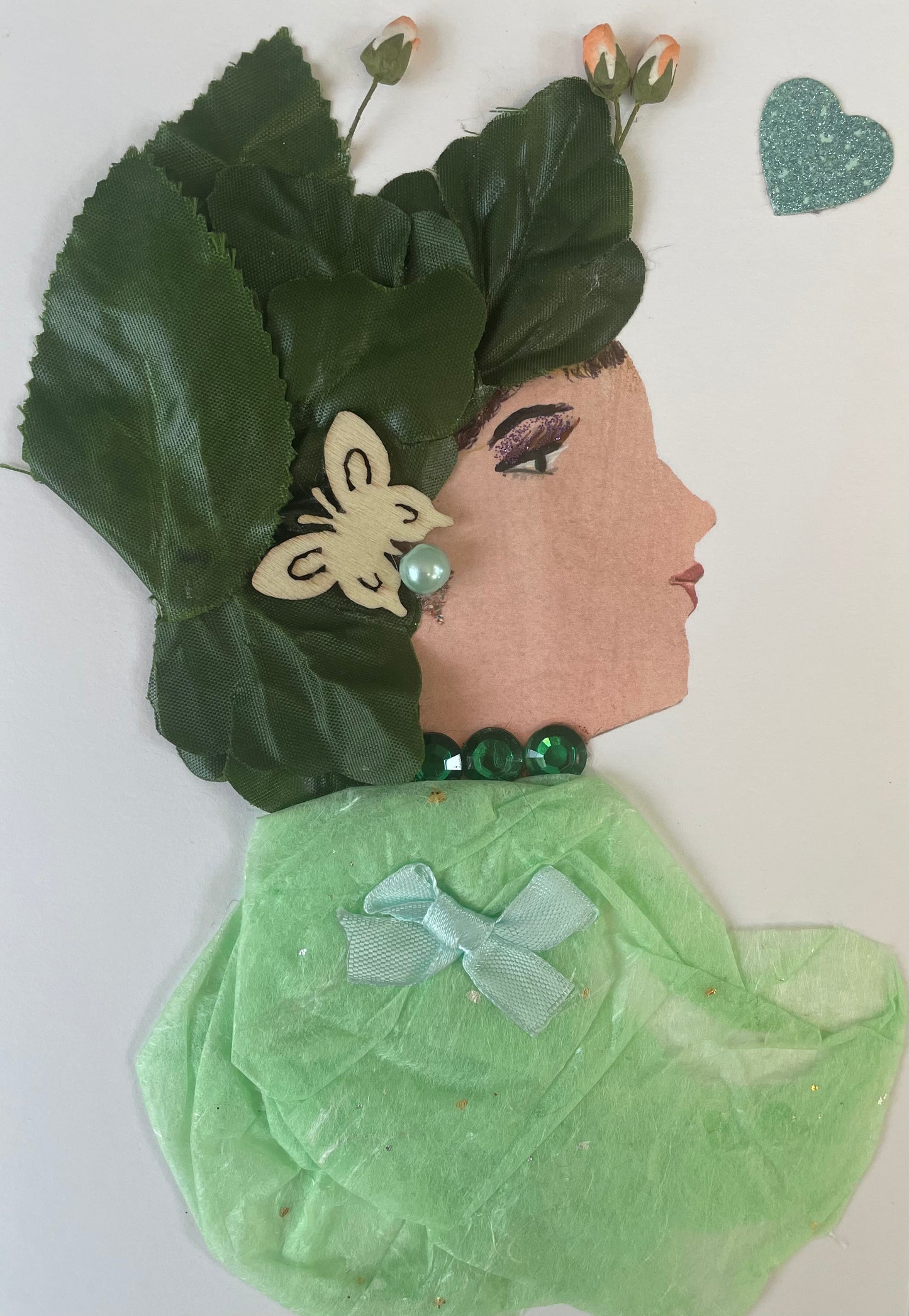 This handmade card called Celery Cyprus is wearing a light green blouse that she pairs it with a light blue bow and dark green necklace. She has a dark green leaf like headpiece with tulips coming out from the top and a butterfly bow.. There is a sparkly blue heart to the right of her. 