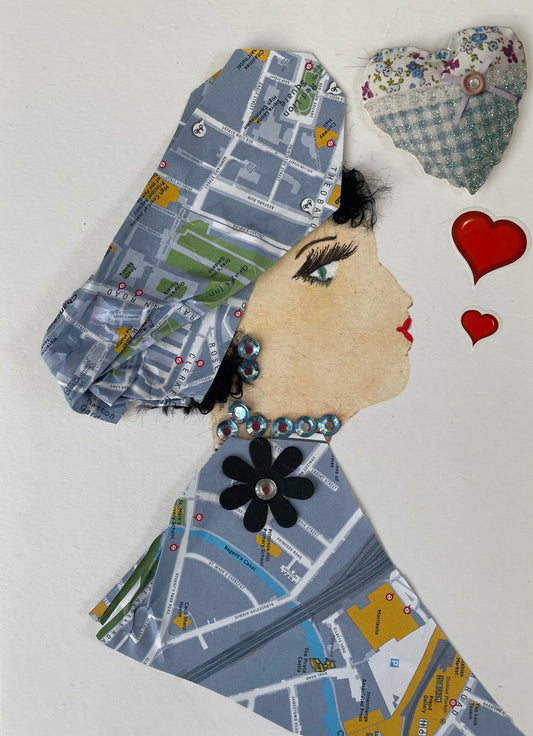 I designed this card of a woman named Blissful Blue. She has a white skin tone  who is wearing a pale blue London map printed blouse with a matching headpiece. She has a black flower on her blouse. Blissful Blue is wearing similar shiny blue jewel-like earrings and necklace. There are 2 red hearts to the right of her with a bigger quilted heart above those.