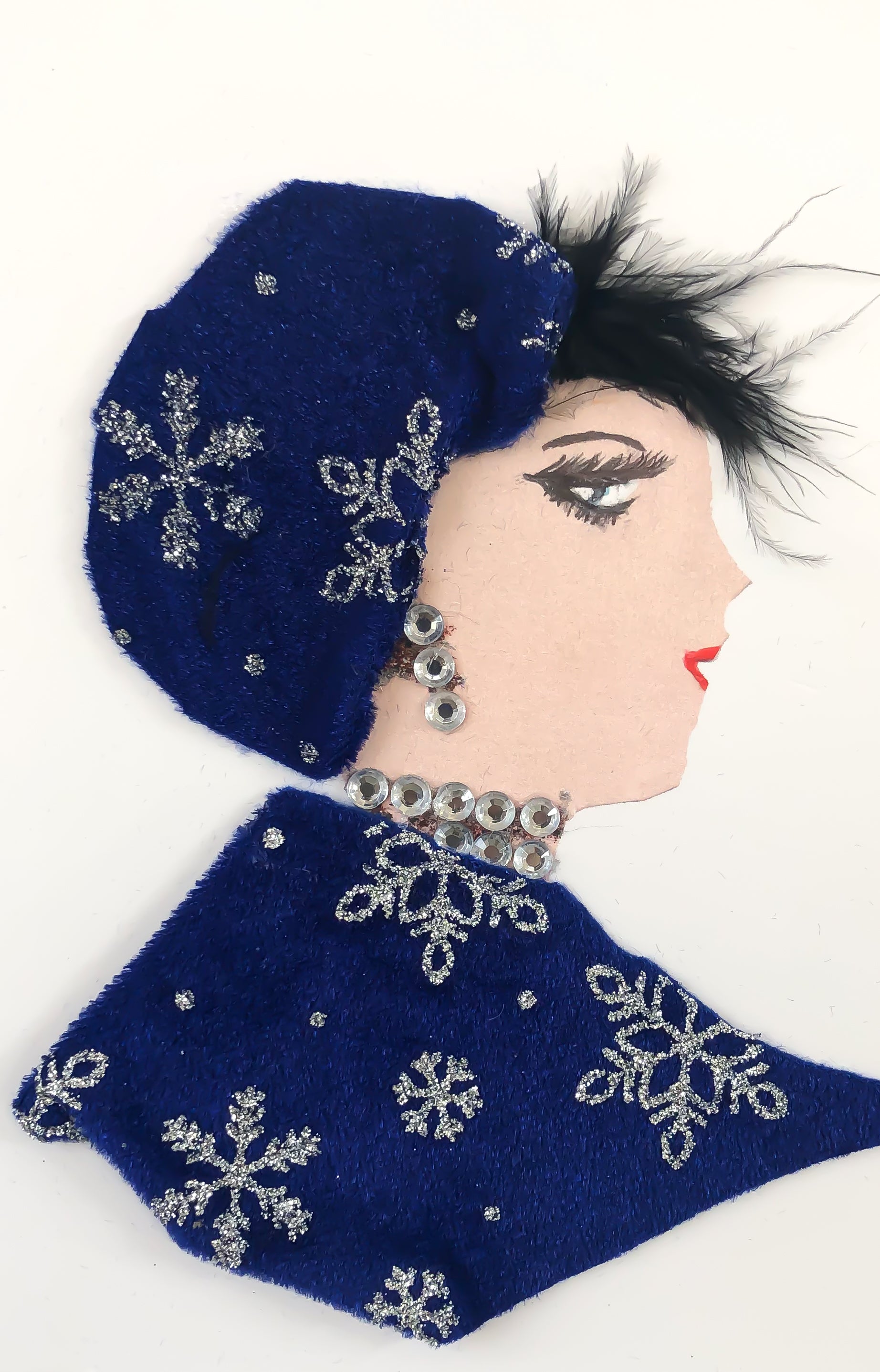 This card is of a woman named Betty Kingsbury. She wears a matching dress and headdress which is a blue felt material. There is a silver glitter snowflake pattern on both pieces. She wears a diamond gem necklace and matching earrings. Her black, feathery hair peaks out of the top of her headdress