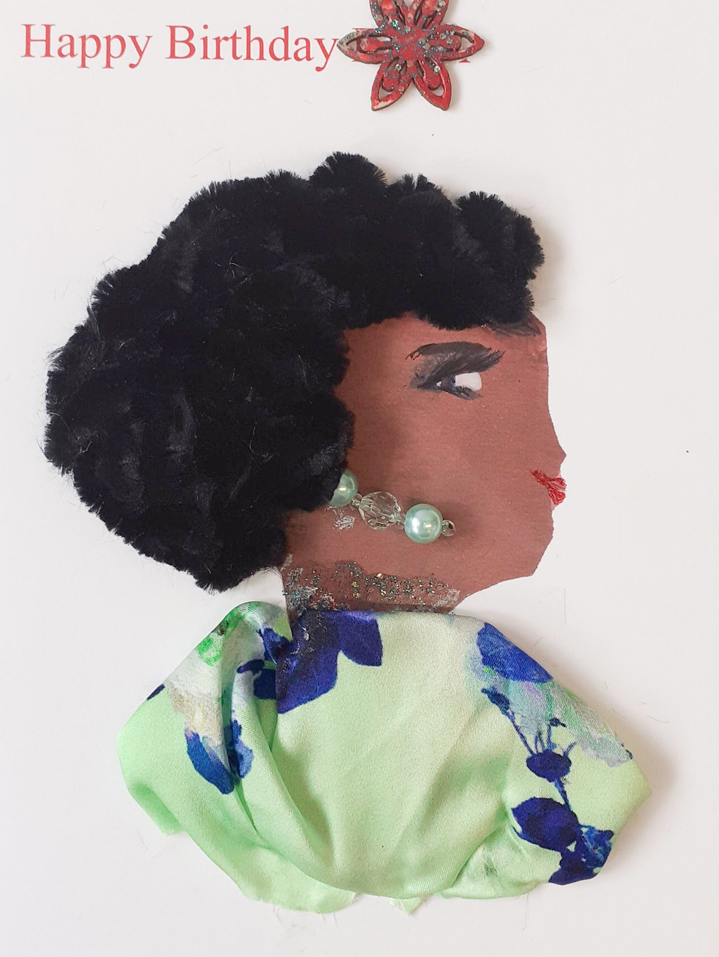 This card displays a woman named Olayinka. She is wearing a light green blouse with a blue plant pattern on it and matching green pearl earrings. Her hair is short, curly, and black, and made of an almost fur like material. On the top left corner, it says happy birthday and there is a red flower next to it. 