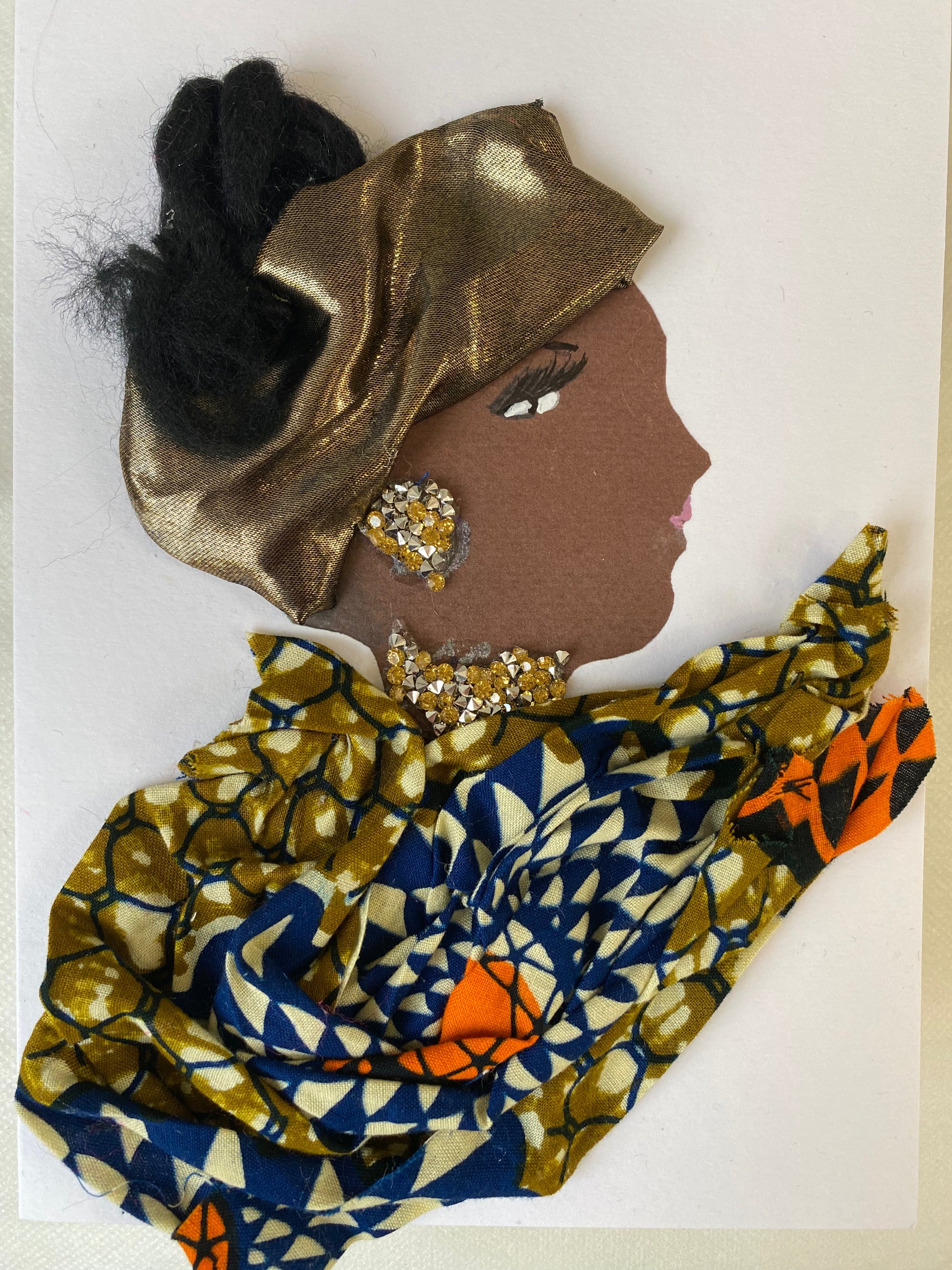 This is a handmade card of a woman named Maybelline Merry. Her hair is styled in soft black fibres, partially wrapped with a shiny gold fabric. She wears sparkling gold gem earrings and a matching necklace. Her outfit is made from patterned fabric in deep blue, olive green, and bright orange, arranged in elegant folds.