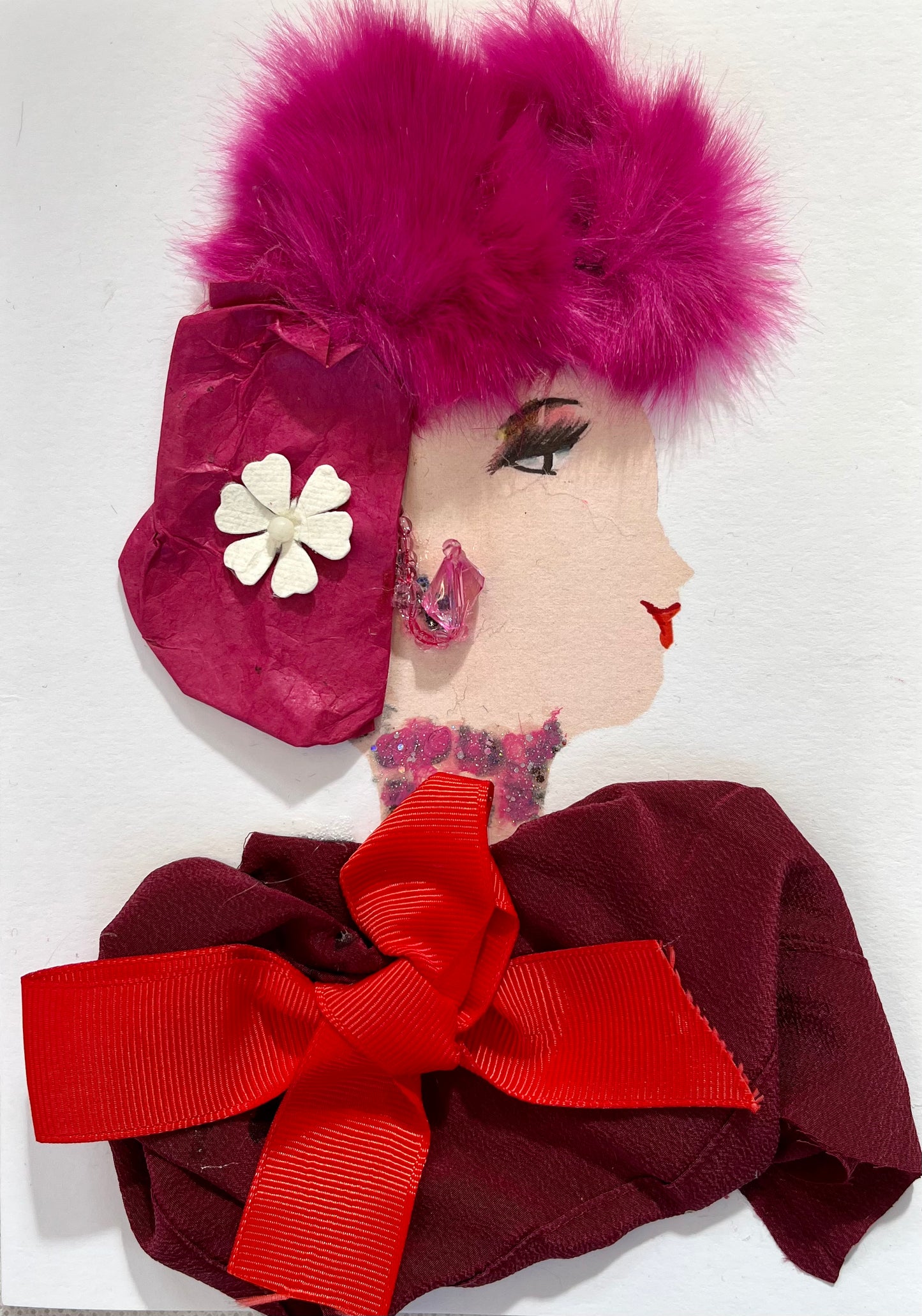 This card is called fuschia. She wears a dark red blouse with a bright red bow. Her hair is bright pink and furry, and she has a headscarf in that is the same colour. It has a white flower on it, and she wears pink jewellery. 