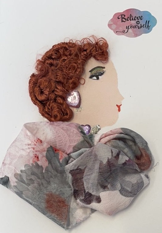 This card depicts a woman wearing a gray and pink floral dress. Her earrings and necklace both have a large pink gem on them. In the background, there is a sticker which says "believe in yourself" 