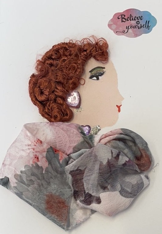 This card depicts a woman wearing a gray and pink floral dress. Her earrings and necklace both have a large pink gem on them. In the background, there is a sticker which says "believe in yourself"