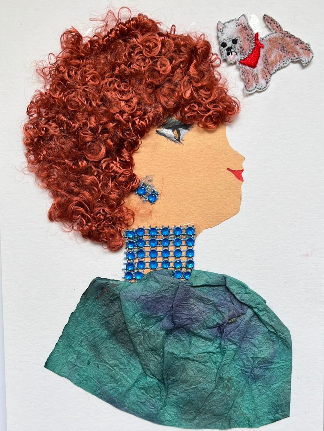 This card has been given the name Bea. Bea wears a blouse which has different tones of blue blended together, and brighter blue gem jewellery. Her hair is red and curly, and it is short. In the top left, there is a small dog with white and ginger fur, and it is wearing a red bandana. 