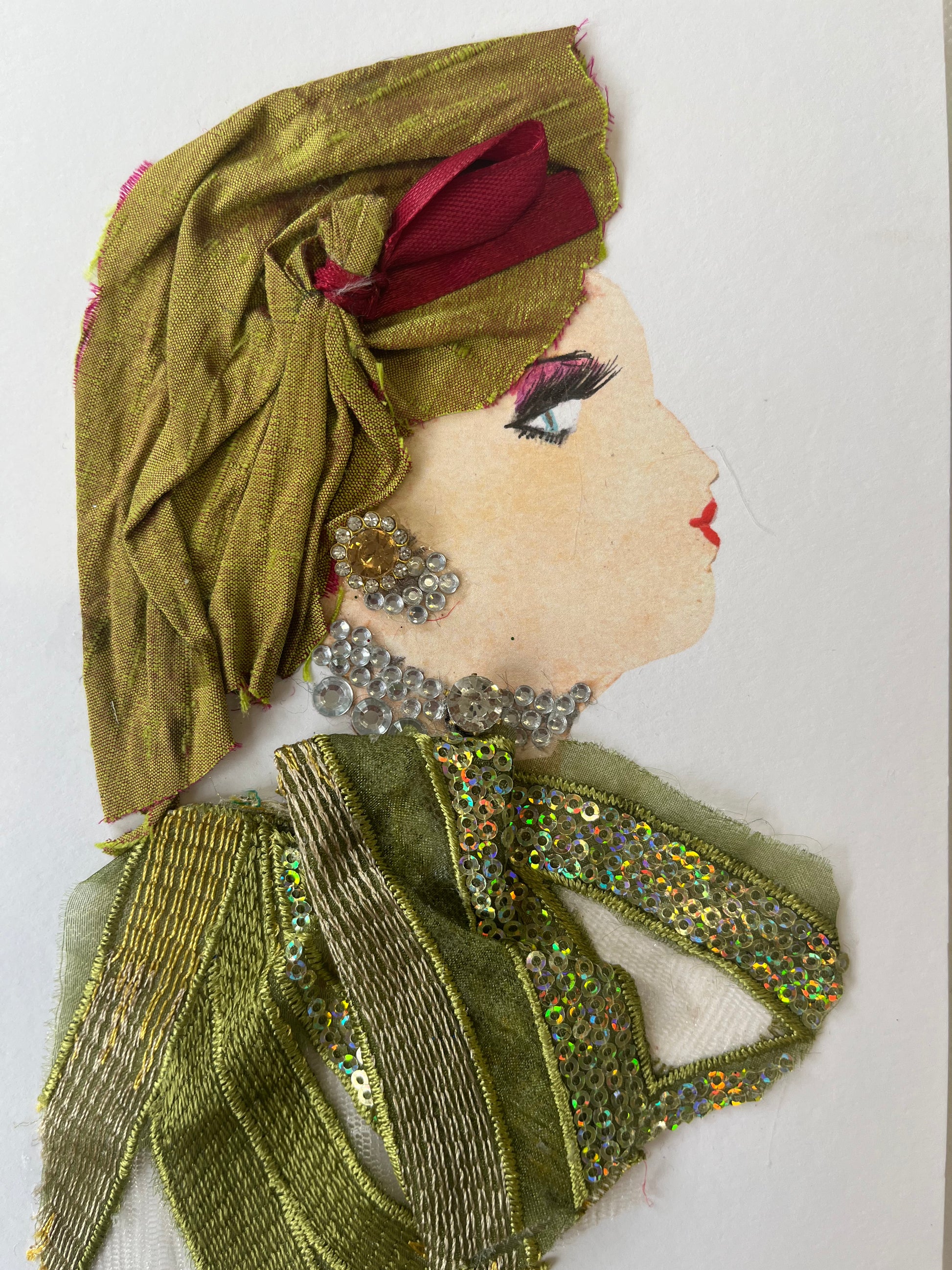 This card has been given the name Sally Sparkles. She wears a top made of ribbons of olive chiffon and sequins. She wears a headscarf that is also olive green but has an accent of rose pink in it. Her jewellery is a large diamanté earring with a yellow stone in the middle, and a matching diamanté necklace. 