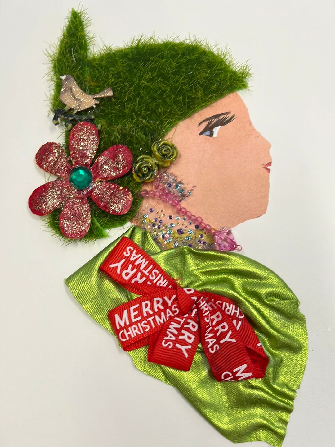 She wears a silk green blouse with a bow that says merry christmas. Her hair is made of a grass fabric. She wears a glittery red flower and a small bird in her hair. 