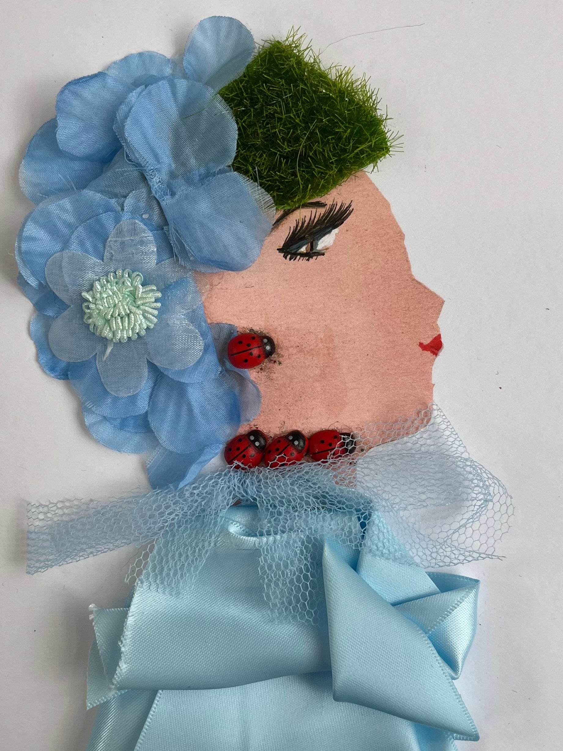 This card has been given the name Gillian Jubilee. Her blouse is a powder blue silk material, and she wears a matching netted scarf. Her hair is made of a grass looking fabric, and she has blue flowers scattered in it. She wears jewellery that looks like ladybugs. 