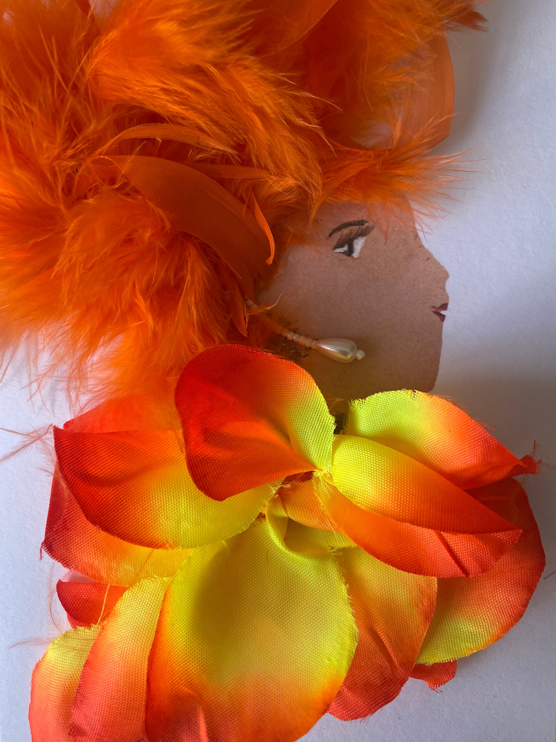 I designed this card of a woman named Magnificent Millie. She has a white skin tone and is wearing an array of electric orange feather in her hair. She wears a blouse full of orange and yellow flower petals. She wears dangle pearl earrings.