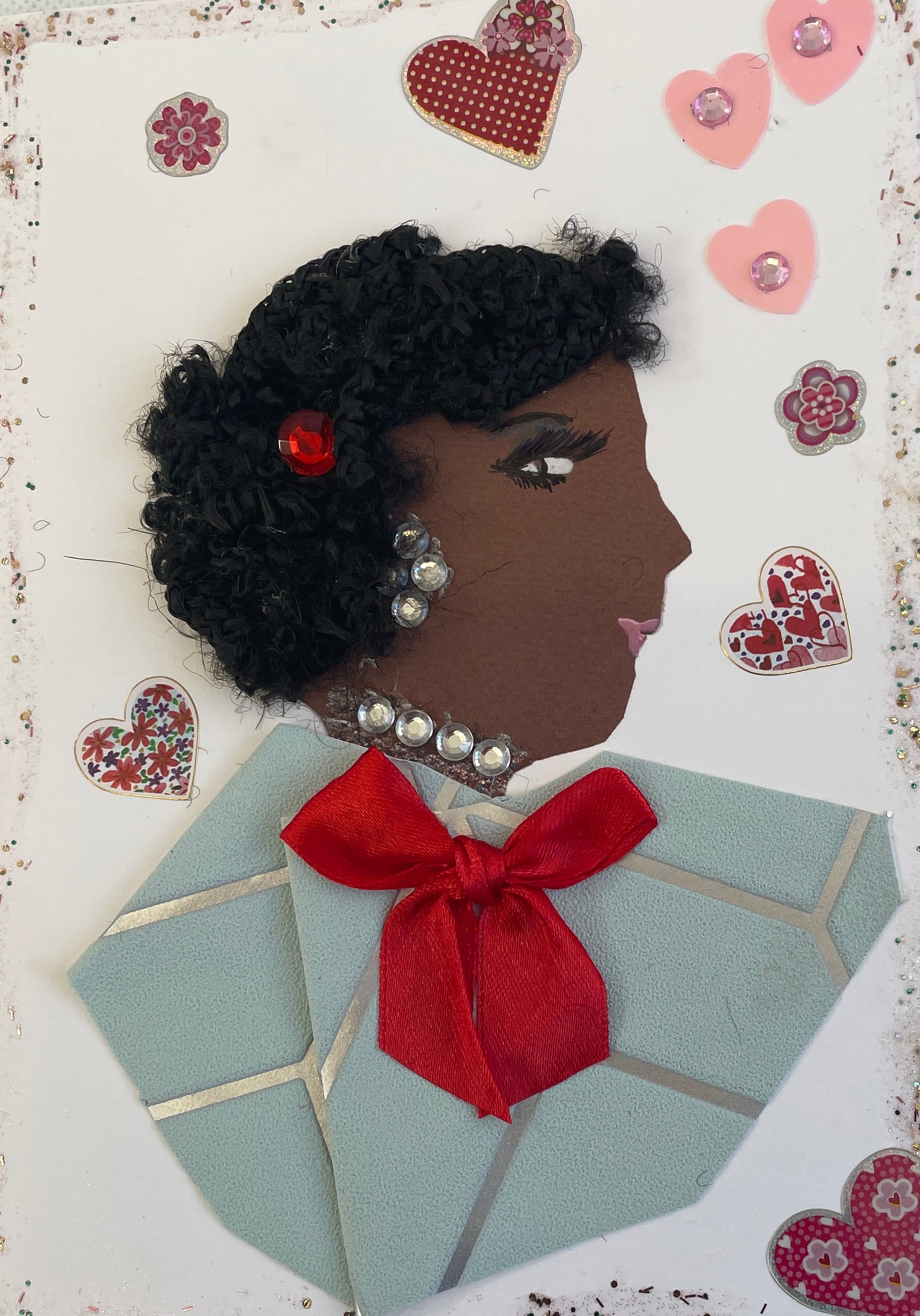 This card is of a woman given the name Happy Hampshire. Happy Hampshire wears a pale blue blouse with a silver line pattern running through and a red bow on the front of it. Her necklace and earrings are both diamanté gems, and there is a red gem in her short, black, curly hair. Surrounding her there are red and pink hearts with various patterns, and a glitter border surrounding the entire card.  