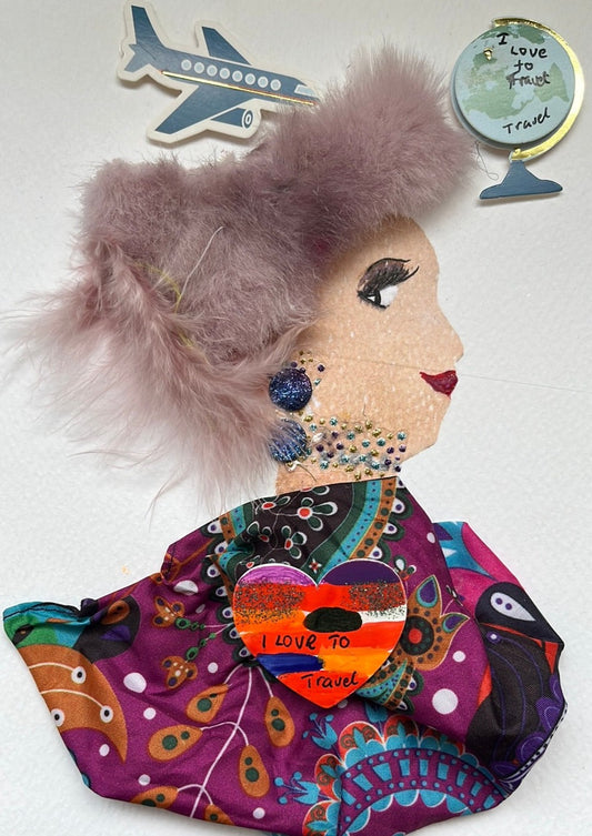 This card has been given the name Sasha. Sasha wears a blouse made of a patterned material with flowers, paisley, and polka dots on it with purple, blue, and yellow accents. Her hat is made of a light purple furry material, and peaking out is her dark blue sparkly earrings. In the top left corner, there is an airplane sticker, and on the right corner, there is a globe that said "I love to travel" on it. 