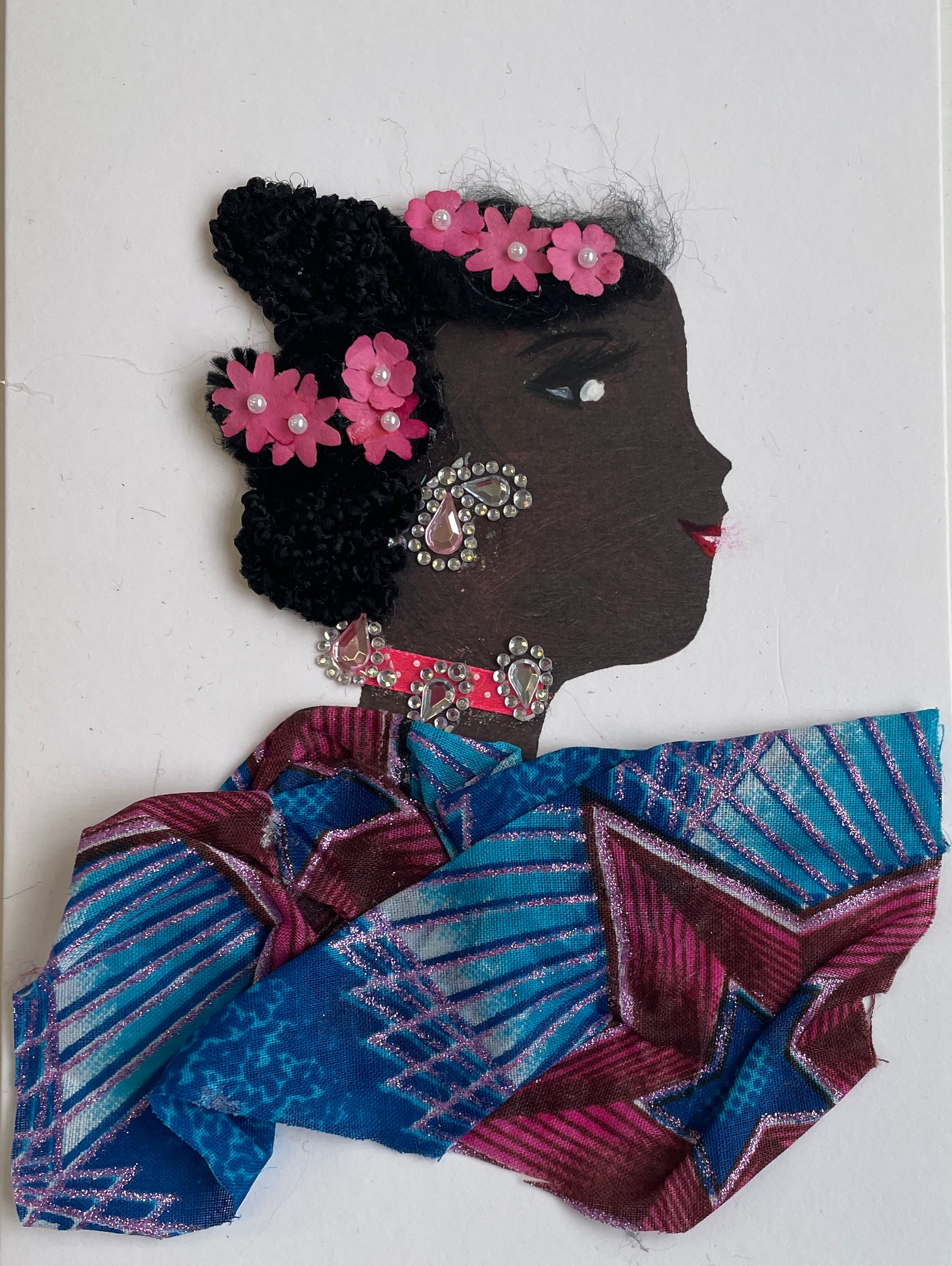 I designed this card of a woman named Shayo Superior. She has a black skin tone who wears pink flowers in her hair. She wears a blue and red pattern blouse. She wears silver and pink jewellery. 