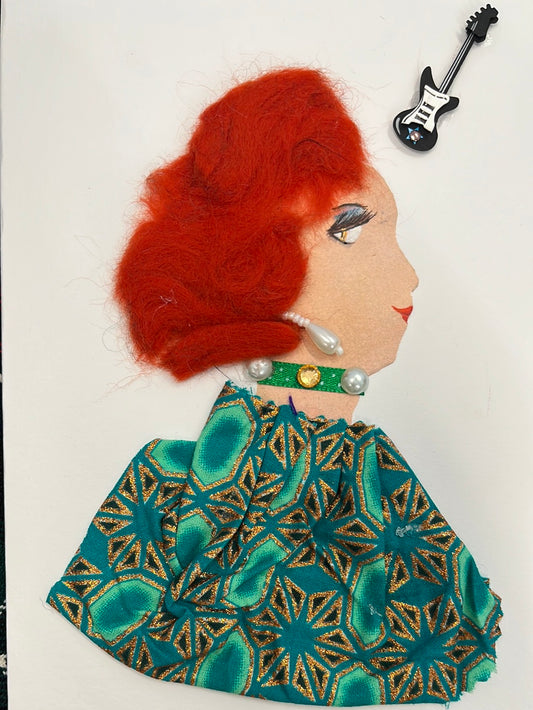 This card is given the name Derby Countess. She wears a green and gold geometric printed fabric as her blouse, and has flaming red short hair. In the top right corner, there is a small sticker of a guitar.