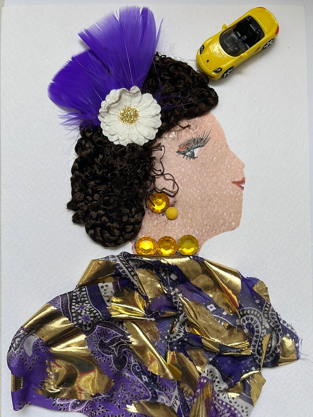 This card was given the name Halima. Halima's blouse is made up of a purple backgrounf with gold paisley accents,  and large gold stripes running through the entire top. She wears a yellow gem necklace and matching earrings. Her hair is deep brown, and has a white flower in it that has a sparkly gold center. Behind the flower there are two purple feathers. In the top right corner, there is a small toy sports car which is light yellow. 