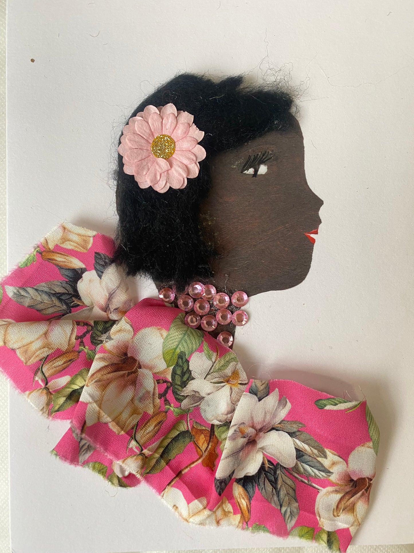 I designed card of a woman named Bella Bakerloo. She has a black skin tone and wears a pink flower in her hair. She wears a pink floral blouse. She wears pink gem jewellery. 