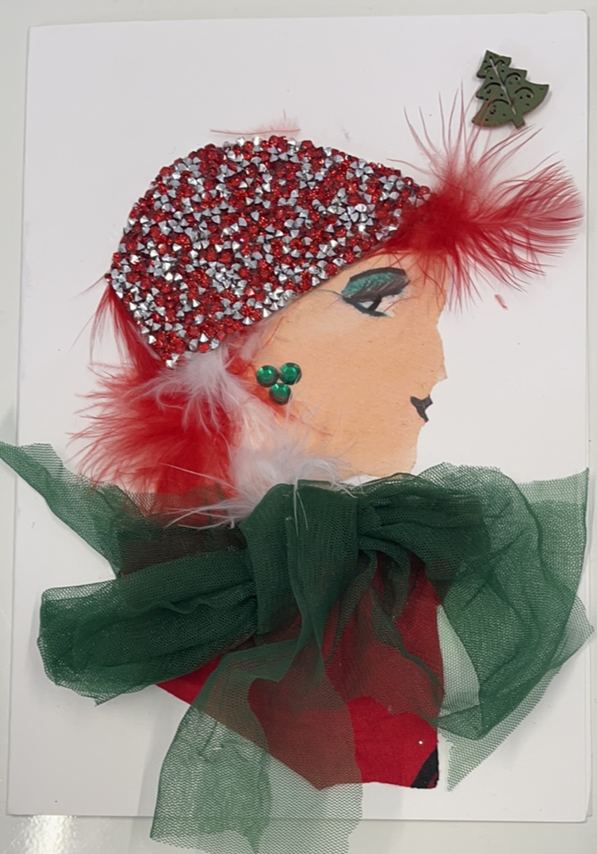 This card is given the name Erinn. She wears a red blouse with a green chiffon bow on it. She wears a hat which is covered in red and silver diamantés, and there are small red and white feathers sticking out of the bottom of it. In the top right corner, there is a small Christmas tree sticker. 