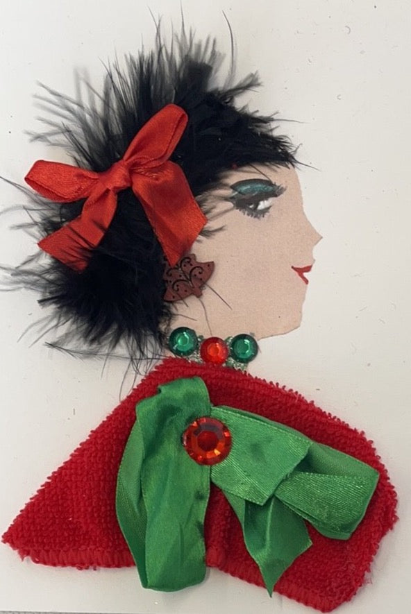 This card has been named Christmas Belinda. She has a white skin tone and she wears a red towel material blouse with a green bow. There are green and  red gems as her necklace. She wears a red satin bow in her black feathery hair. 