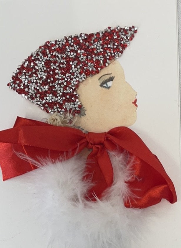 This card has been given the name London Angel. She wears a white feathery blouse with a large red bow around the neck. She wears a red and silver diamanté headdress and her blonde curly hair peeps out of the bottom.