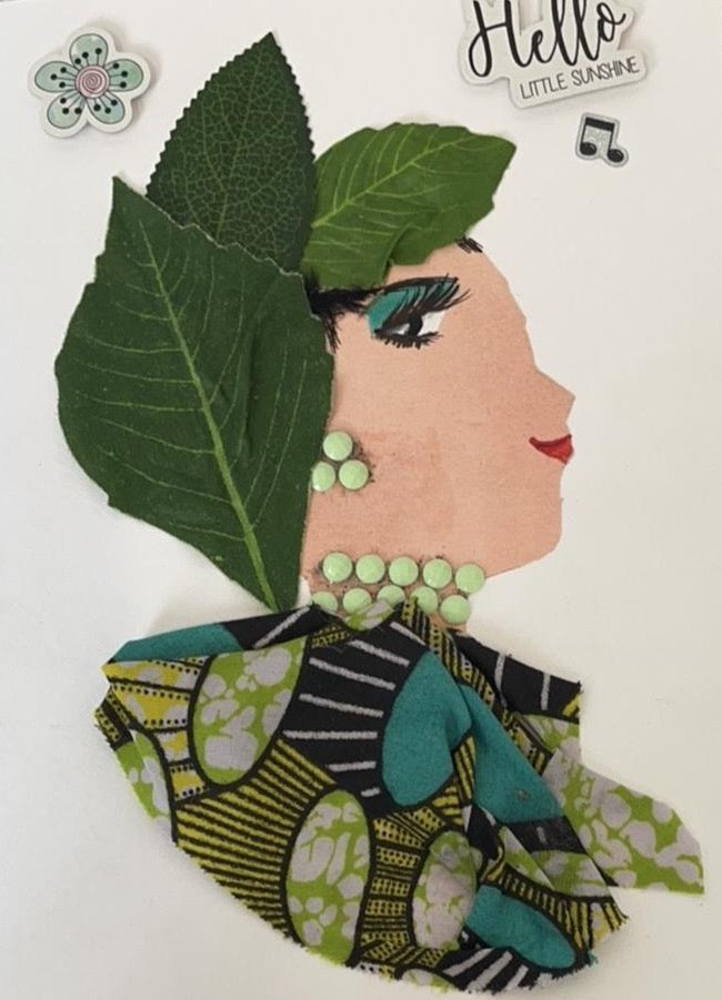 This card is of a woman given the name Greenford Gabi. Greenford wears a printed blouse with green and turquoise circles and yellow and black lines throughout. Her headdress is made of three dark green leaves, and her jewellery is a pale green. In the top left corner there is a small green flower sticker, and in the top right there is a small sticker that says Hello Little Sunshine with a music note. 