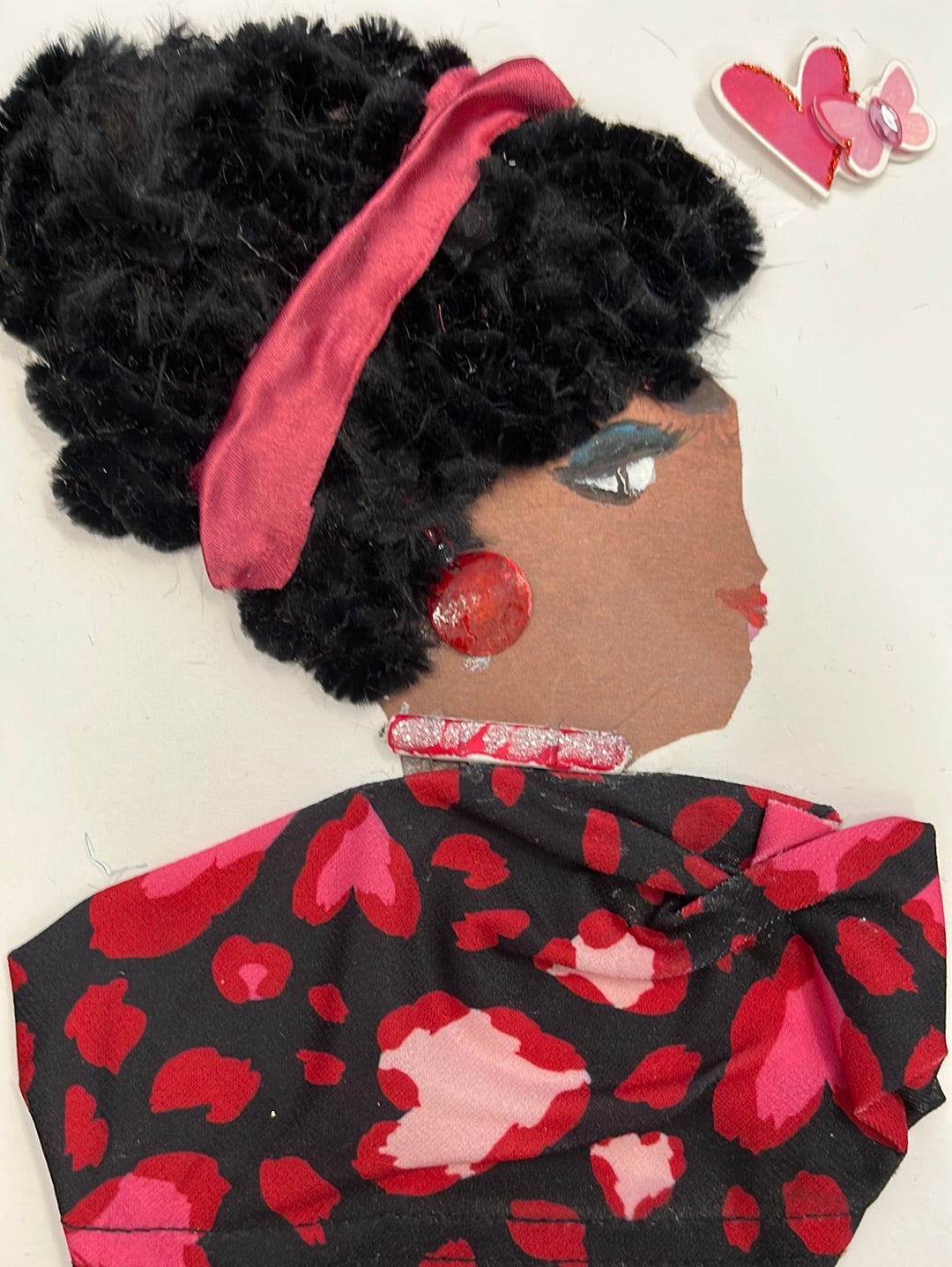 This card has been given the name Miss Maida. She wears a black blouse which has a deep pink cheetah print on it. Her necklace is pink and glittery, and her earring is red. In her black furry hair, she wears a red silky ribbon as a headband. In the top right corner of the card, there is a small sticker of a heart and a butterfly.