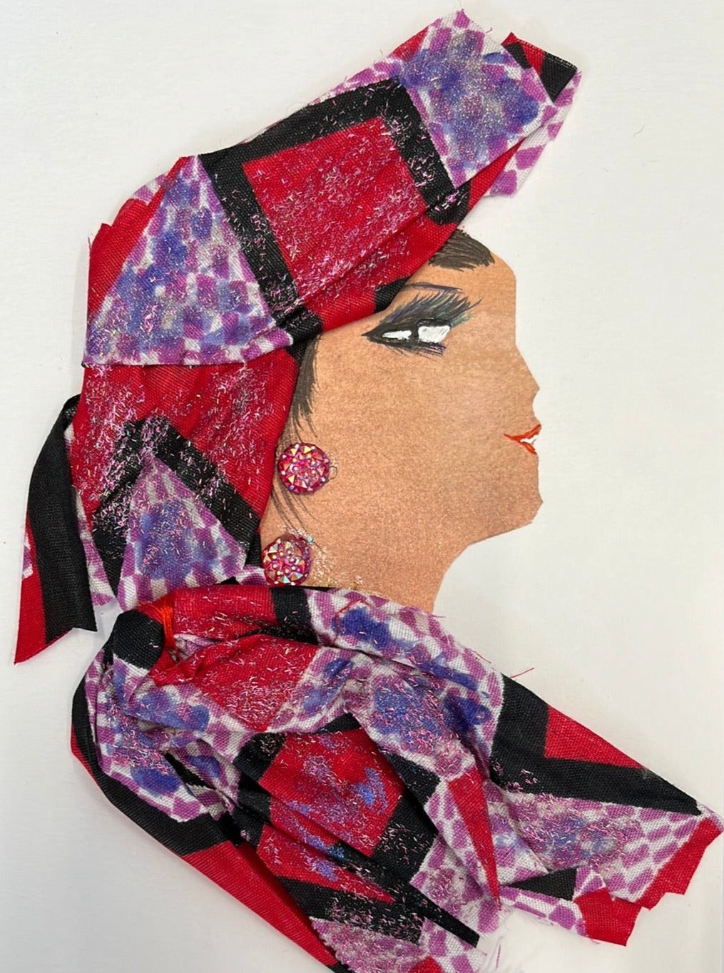 I designed this handmade card of a woman named Ruby Tuby. She has a white skin tone and wears a matching blouse and headdress, which has a red, purple, and black geometric pattern on it and glitter over the entire fabric. Her jewellery is two small red gems. 